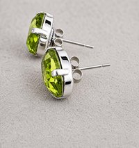 Oval peridot green crystal stud earrings in silver plated prong settings displayed on a neutral fabric background.