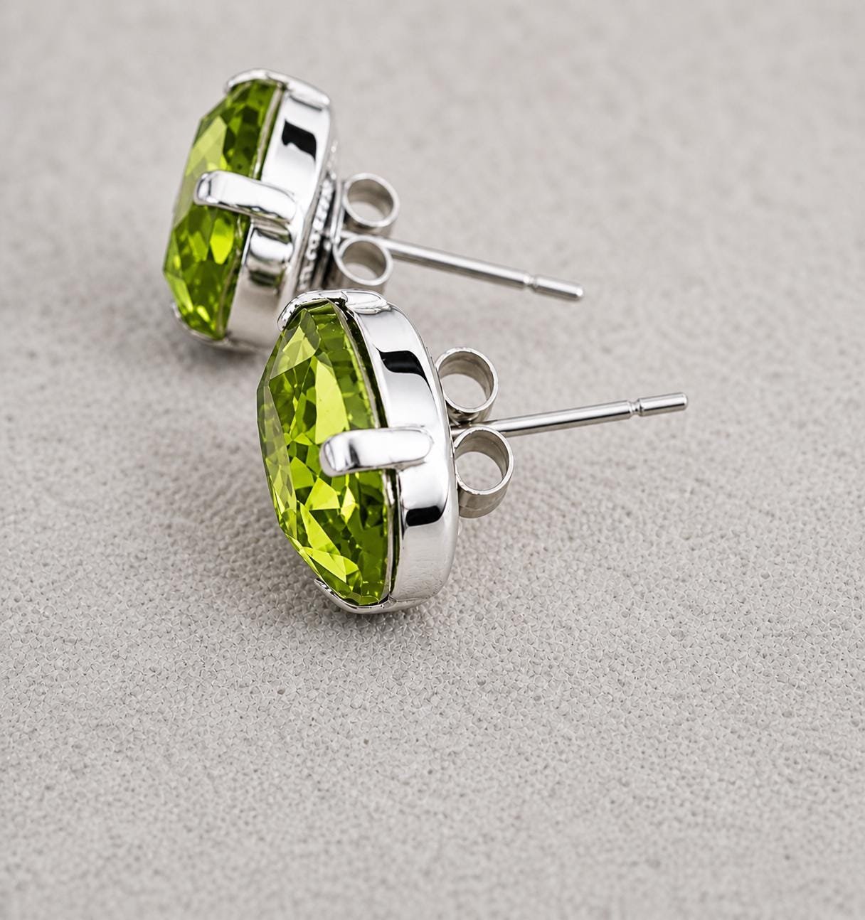 Oval peridot green crystal stud earrings in silver plated prong settings displayed on a neutral fabric background.